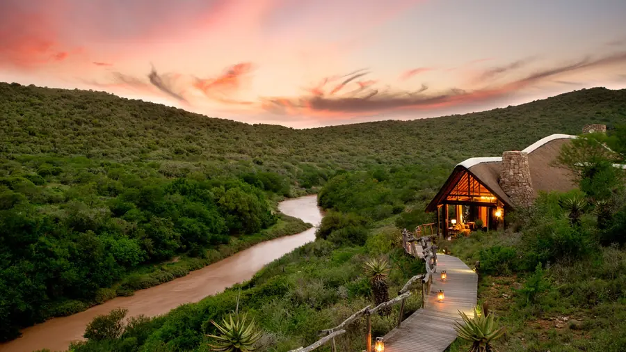 Lantern-lit boardwalk leading to a thatched lodge above a river at sunset in an Eastern Cape malaria-free game reserve, South Africa