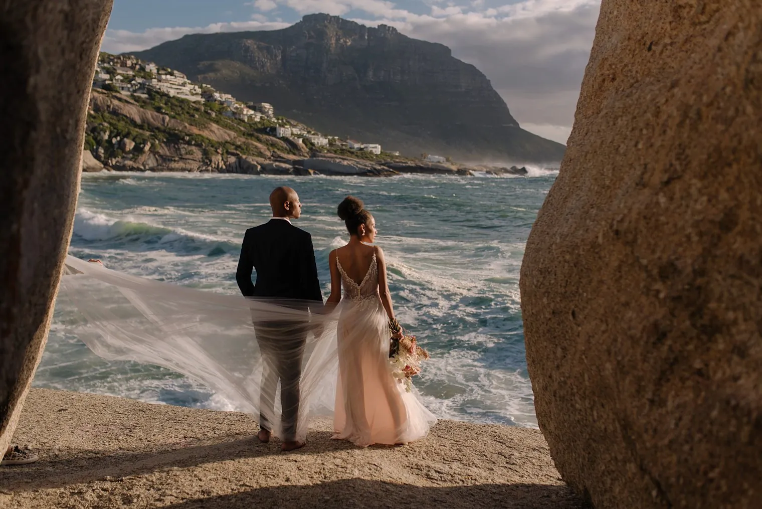 Bride and groom on the rocks above the Atlantic with Cape Town mountains behind