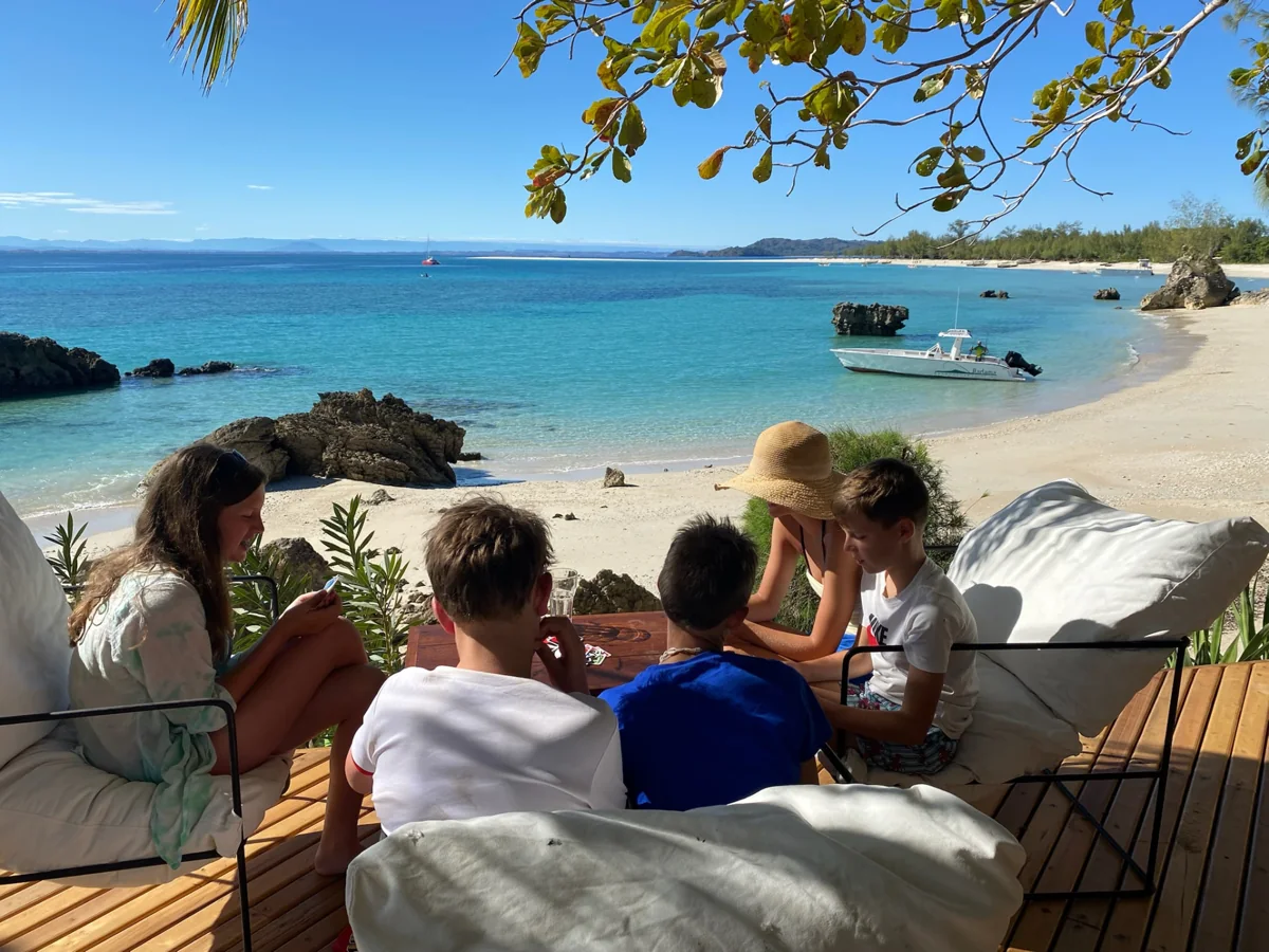 A family relaxing together on a shaded beach terrace beside turquoise water and a tropical sandy shore.
