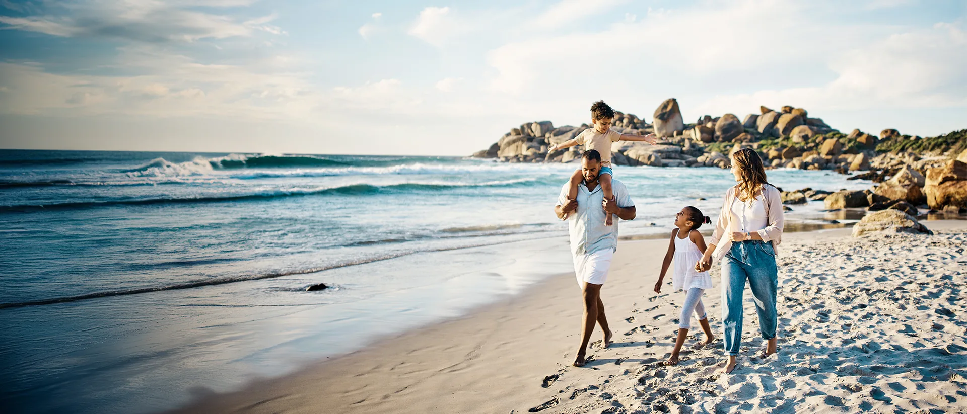 Family walking barefoot along the beach, a boy on his father's shoulders, with surf and rounded boulders behind