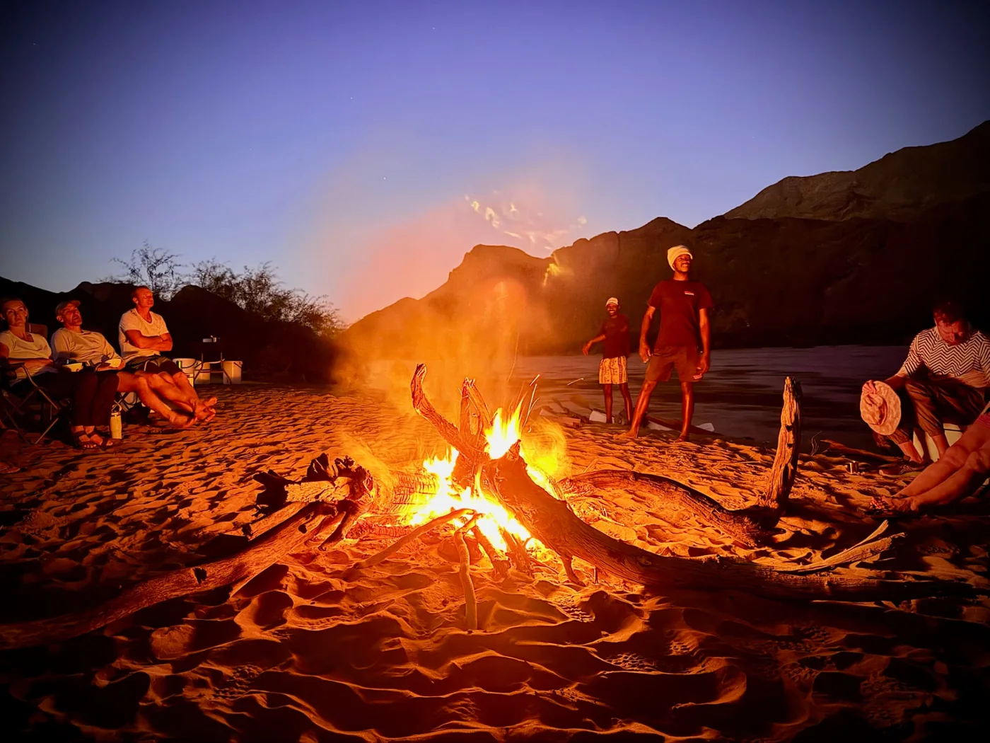 Families gathered around a bright campfire on the riverbank at dusk
