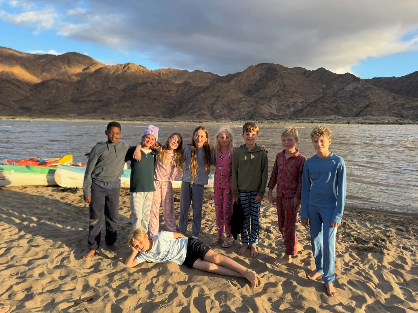 Children posing together on a sandy riverbank in front of canoes and mountains