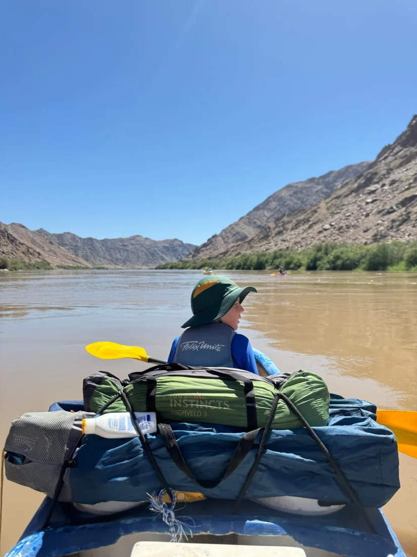 A child in a life jacket at the front of a loaded canoe in a mountain-lined canyon