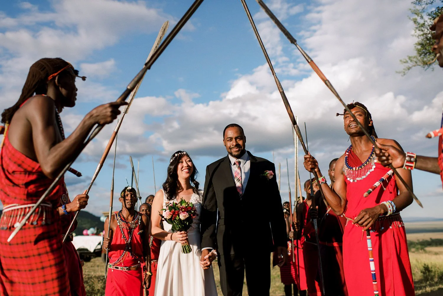 Bride and groom walking through an arch of Maasai warriors' spears on the plains of the Masai Mara