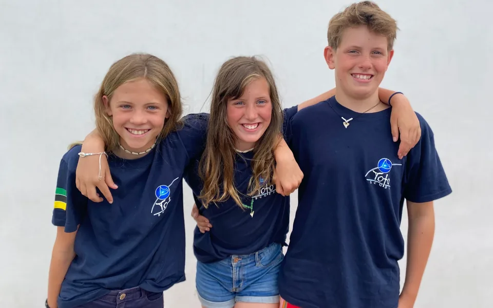 Three children in matching navy Toto Scuba t-shirts standing arm in arm, one with a Tanzania flag patch on her sleeve