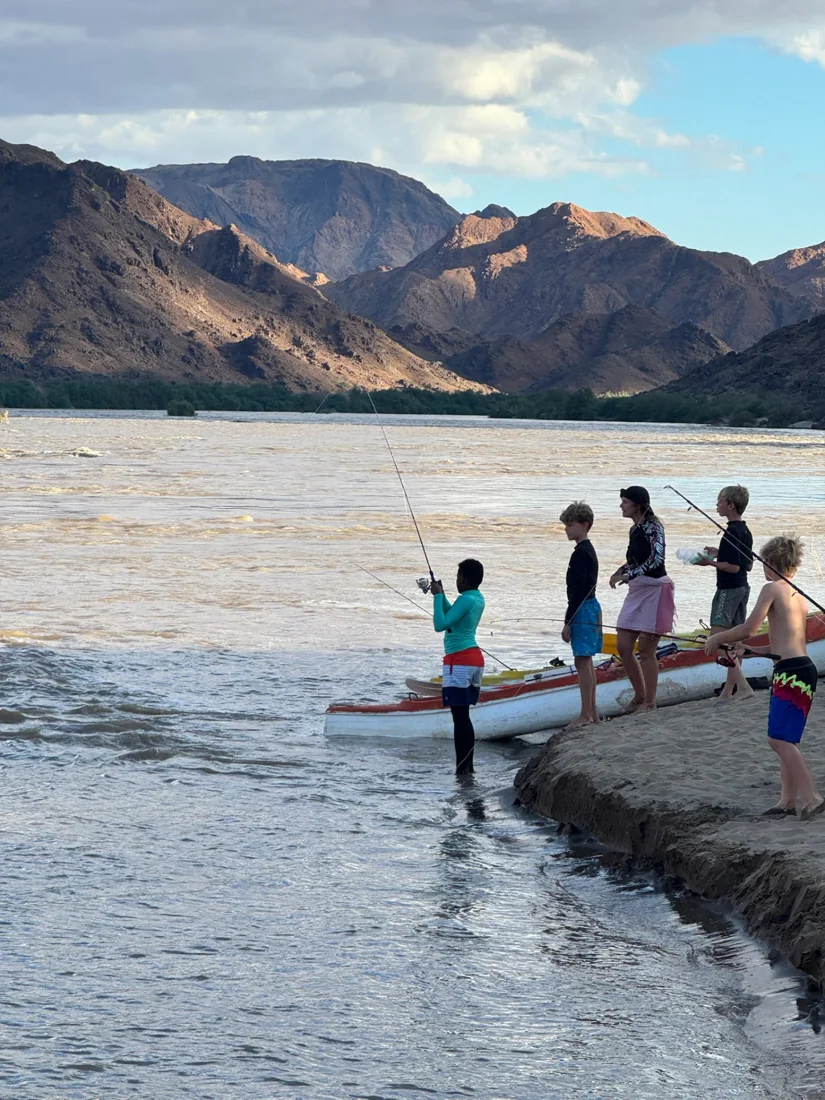 Children fishing from a sandy bank with canoes and mountains behind them