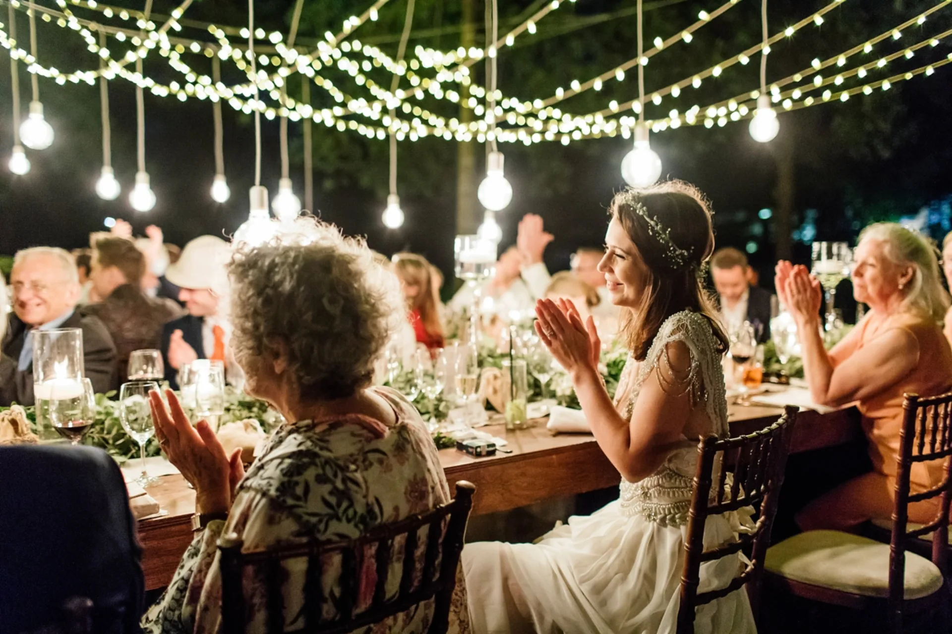 Bride and guests applauding at an outdoor reception under a canopy of fairy lights