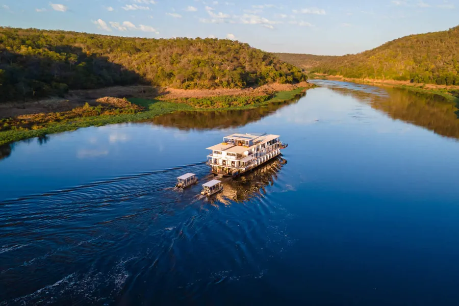 Safari houseboat cruising the calm blue water of Lake Kariba between forested hills at sunset, Zimbabwe