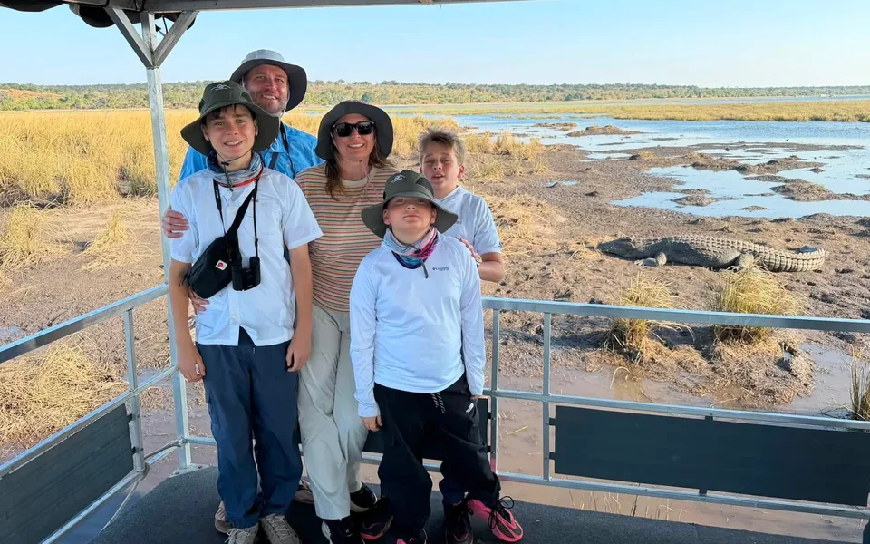 The Baker family of five standing on a safari boat deck with a crocodile resting on the riverbank behind them
